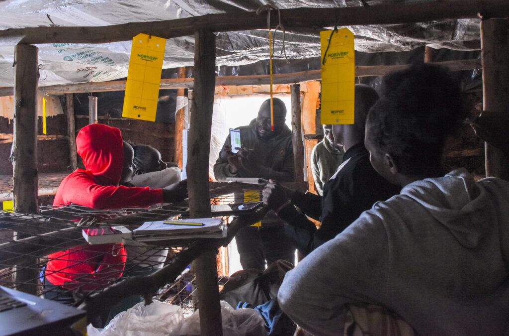 A group of young farmers in Kenya gathered in a hut with a low-ceiling and dim lighting.