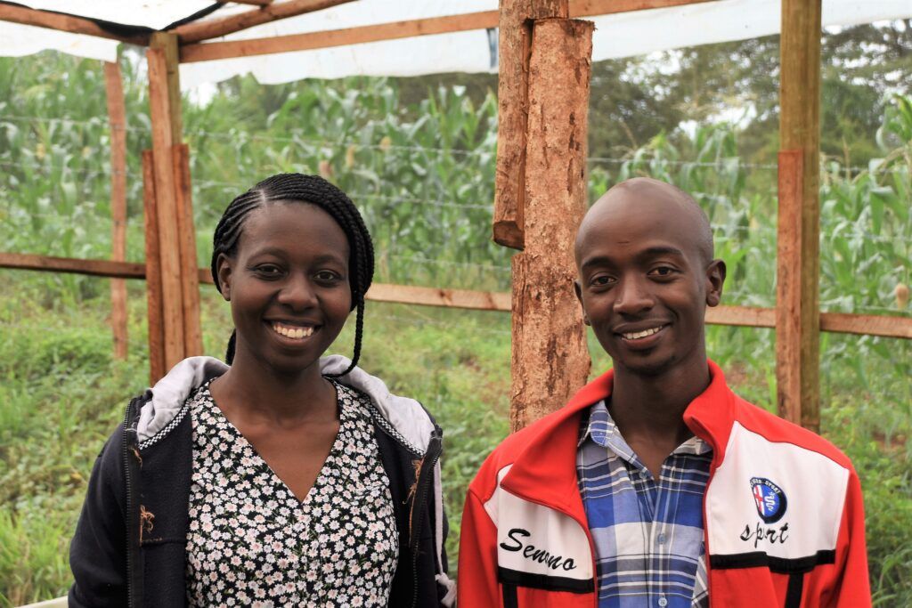 Two young farmers in Kenya, a man and a woman, smiling inside a greenhouse with lush plants behind them.