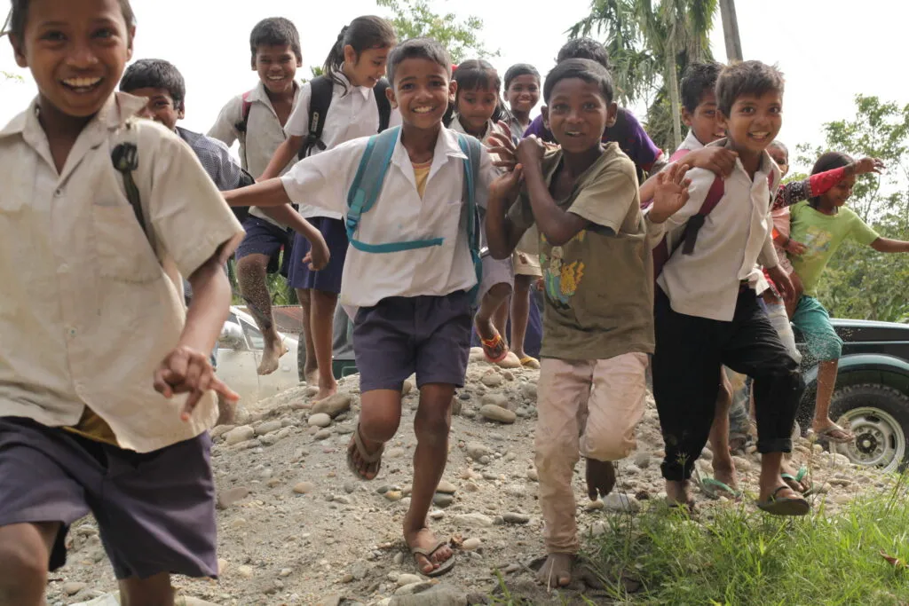 group of smiling schoolchildren run down a dirt road in Assam, India