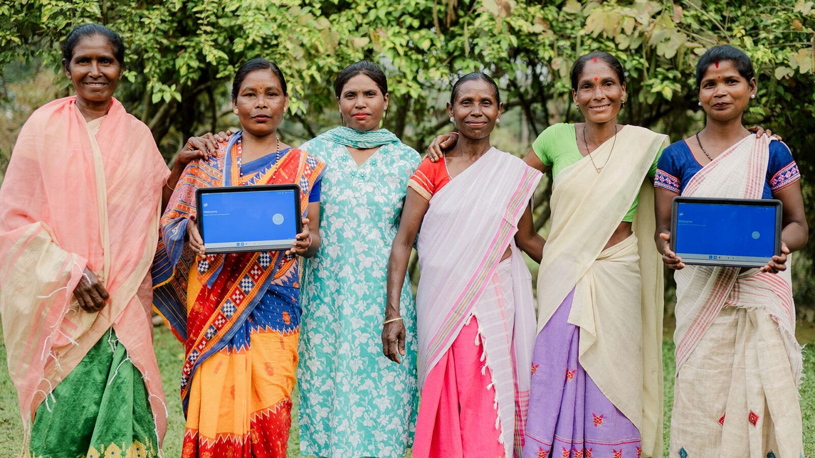A group of women farm workers from India stands in a field. Two of them are holding a tablet displaying the Rainforest Alliance's new gamified training.