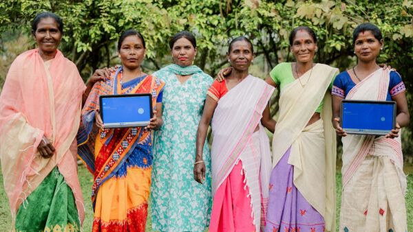 A group of women farm workers from India stands in a field. Two of them are holding a tablet displaying the Rainforest Alliance's new gamified training.