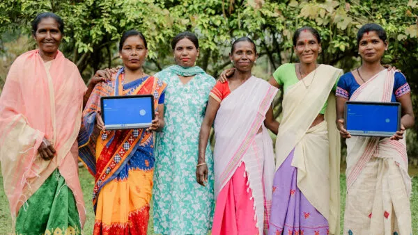 A group of women farm workers from India stands in a field. Two of them are holding a tablet displaying the Rainforest Alliance's new gamified training.