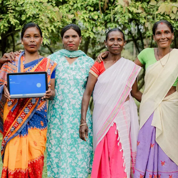 A group of women farm workers from India stands in a field. Two of them are holding a tablet displaying the Rainforest Alliance's new gamified training.